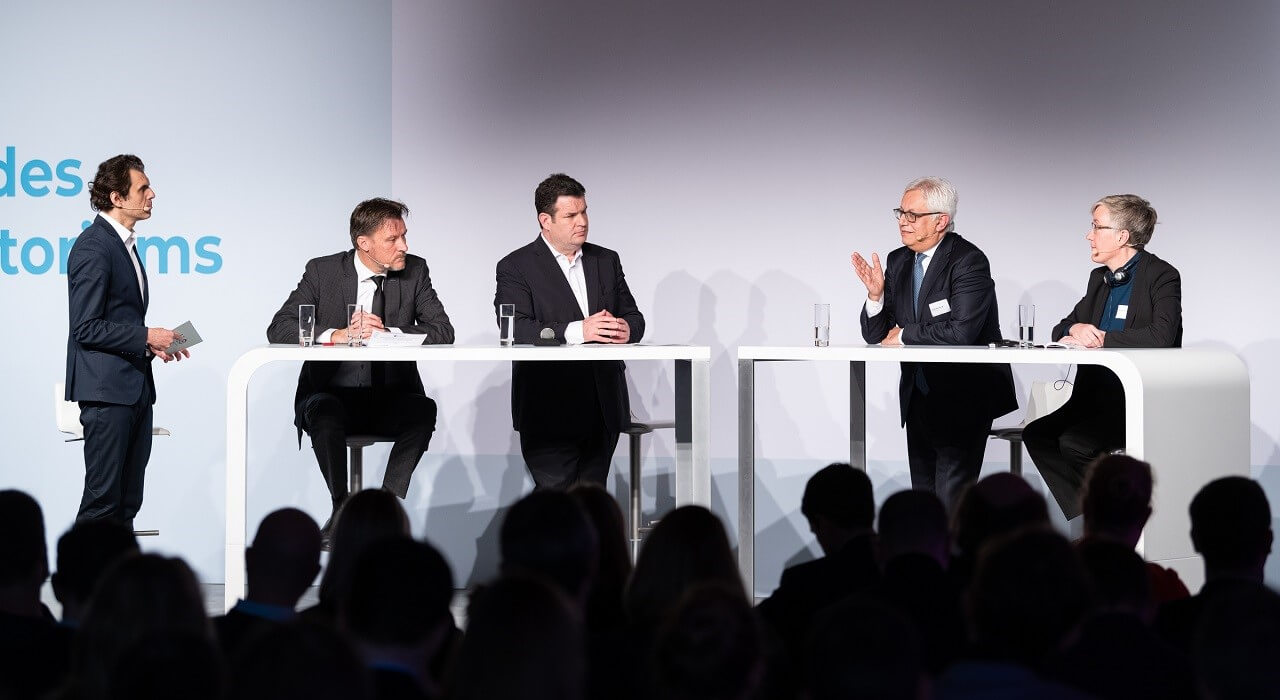 Fünf Personen sitzen oder stehen an zwei länglichen, weißen Tischen auf der Bühne.